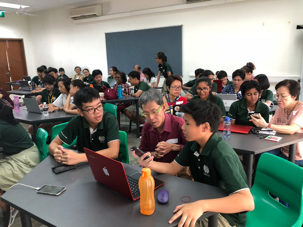 Two students in green uniforms help an elderly man with a laptop in a busy classroom filled with adults and children.